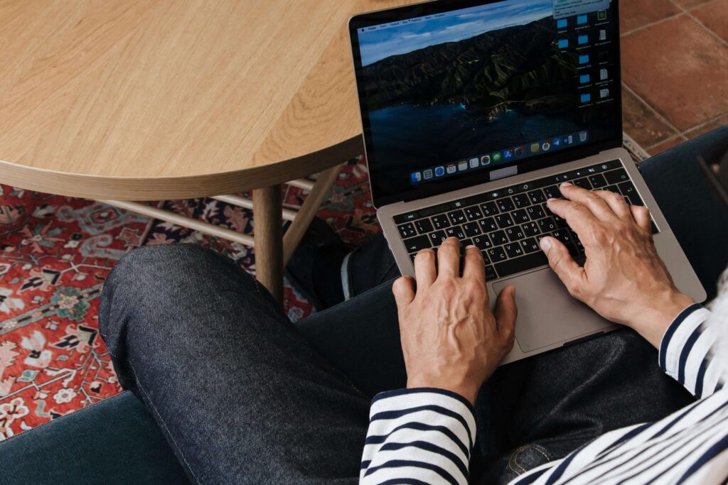 Person working on a laptop while sitting on a cozy couch, surrounded by a wooden table and a patterned rug, illustrating a comfortable work environment