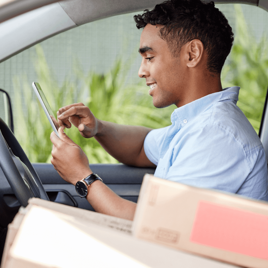 A person sitting in the driver's seat of a vehicle, using a tablet device, surrounded by cardboard boxes representing delivery driver.