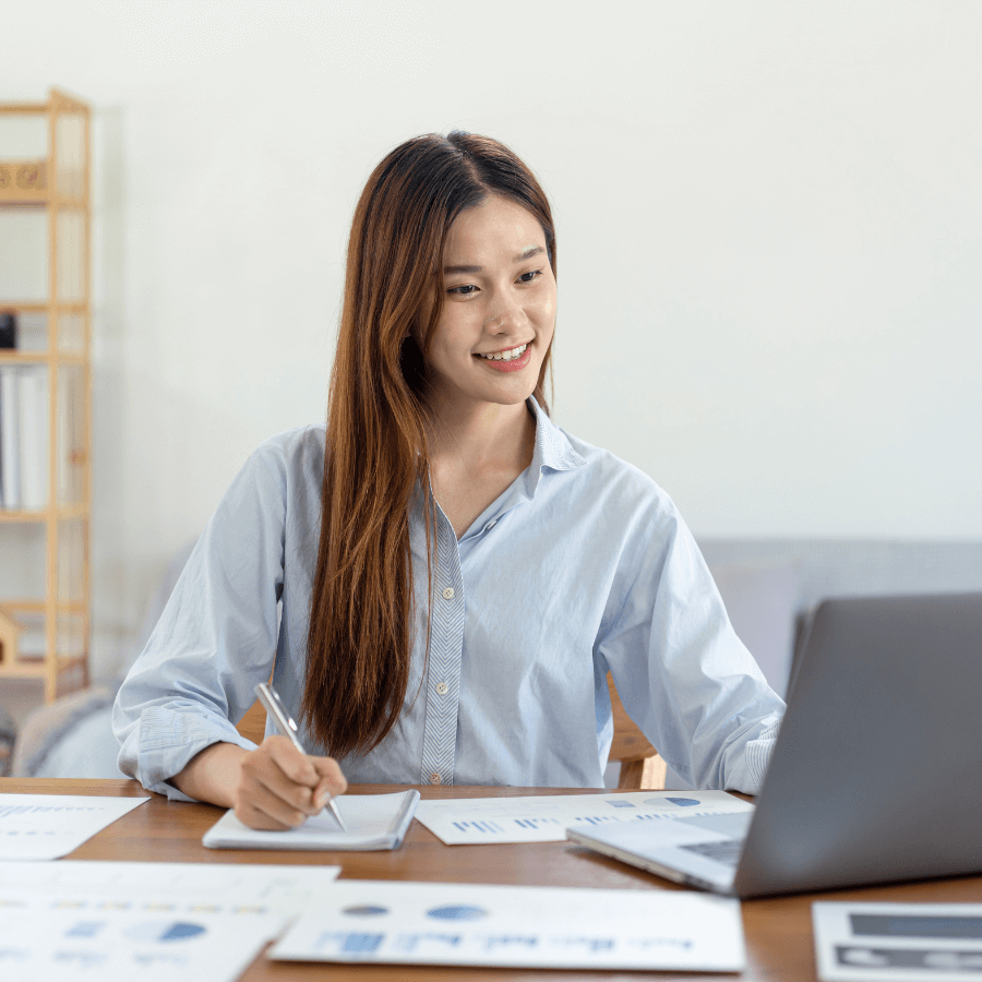 Person sitting at a desk with a laptop, writing in a notebook with charts and graphs on the desk.