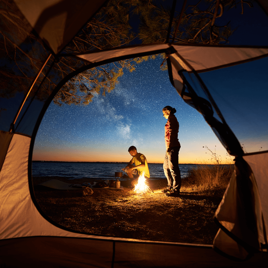 Camping scene at night, viewed from inside a tent, with two people near a campfire under a starry sky.