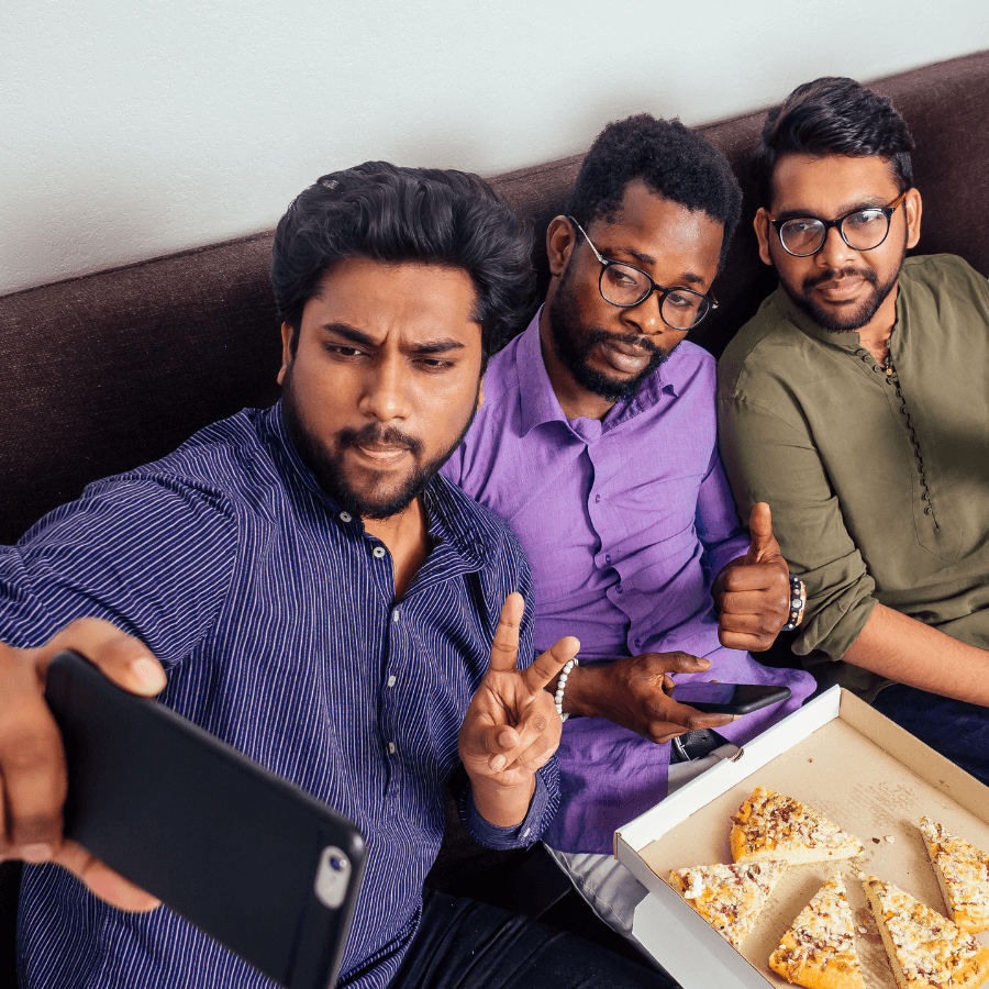 Three friends sitting on a couch, taking a selfie with a pizza box in front of them, enjoying a casual hangout.