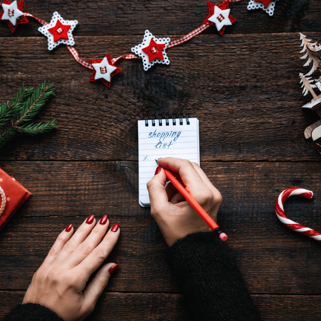 A person writing a Christmas shopping list on a small notepad with a red pencil, surrounded by festive decorations, including a candy cane, star ornaments, a pine branch, and a wrapped gift.