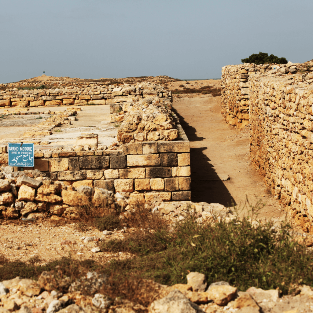 Ruins of the Indus Valley Civilization, showcasing ancient architecture with stone walls and pathways, surrounded by an arid landscape with sparse vegetation and a clear sky.