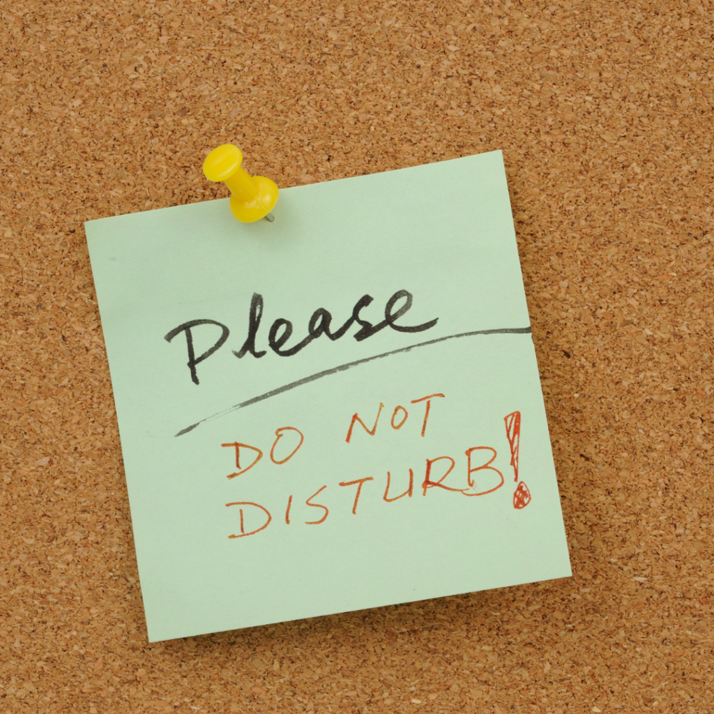 A corkboard with a green sticky note pinned to it using a yellow pushpin. The sticky note has handwritten text that reads: 'Please DO NOT DISTURB!' The word 'Please' is written in black ink, while 'DO NOT DISTURB!' is written in red ink and underlined.